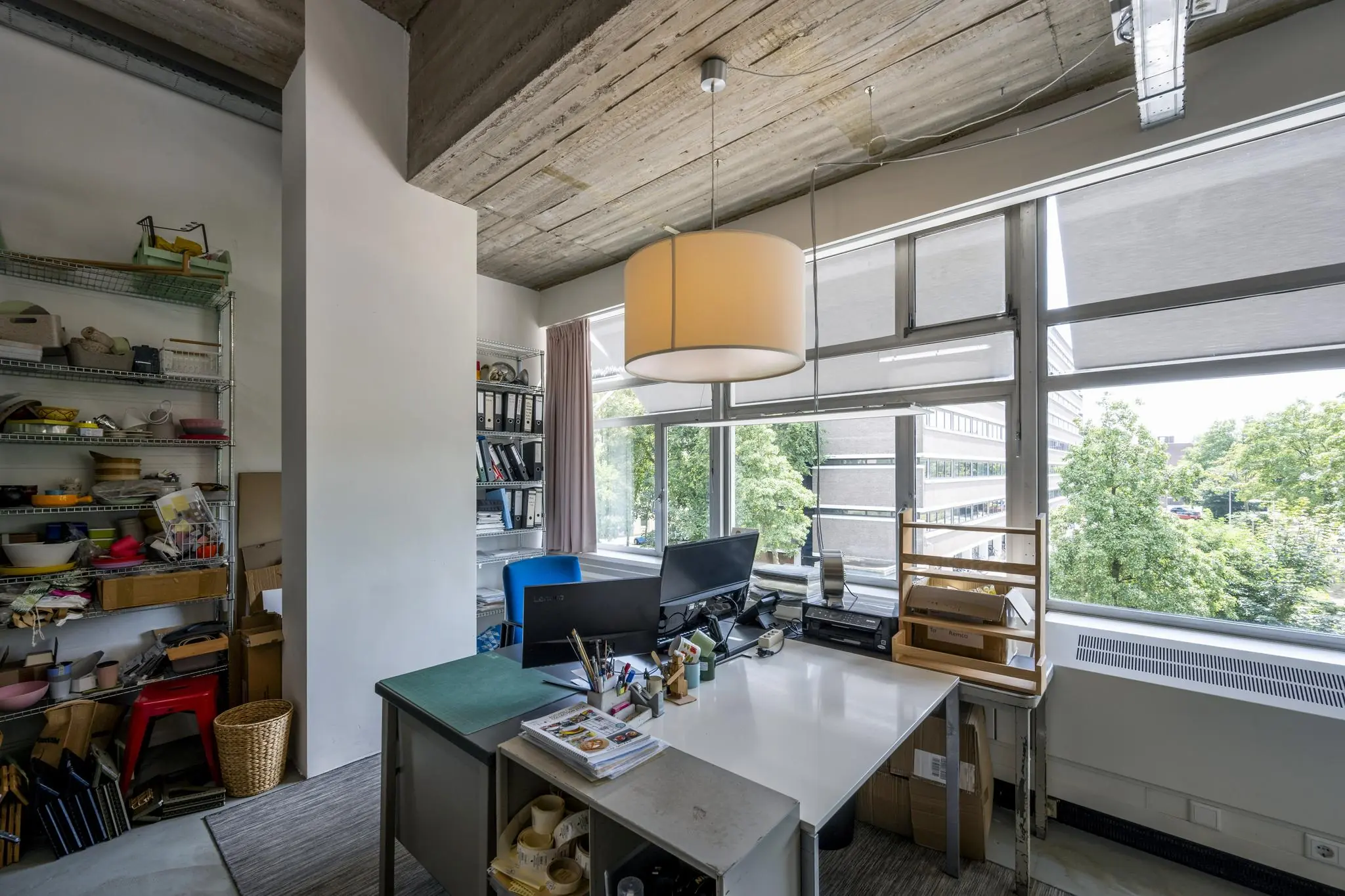 Kantoorruimte aan de Van Leijenberghlaan met groot raam, bureau, opbergrekken en uitzicht op groene bomen.